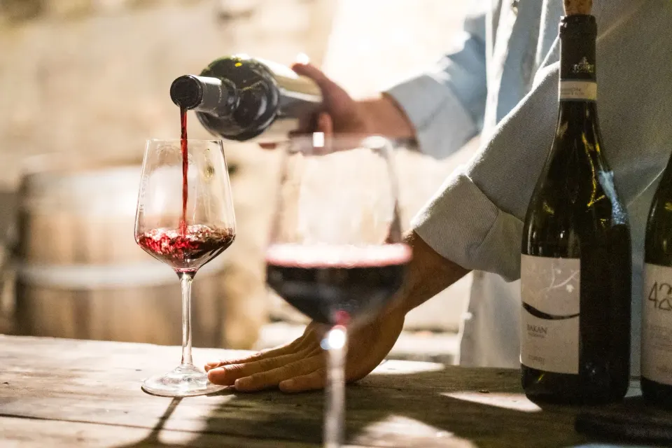 Red wine being poured into a glass on a wooden table, bottles beside it